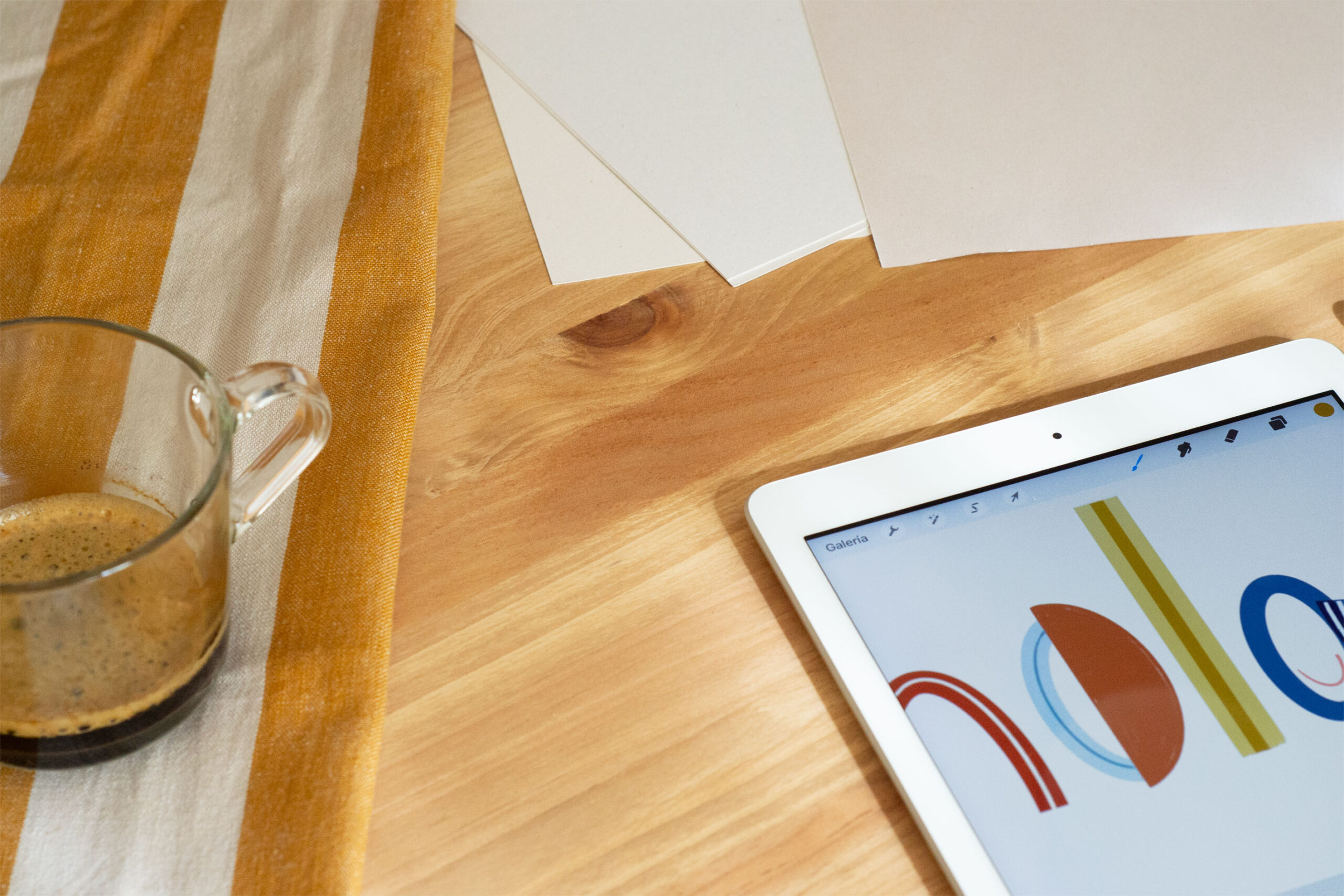 Imagen de la mesa del estudio con un café y un diseño en el Ipad que dice hola